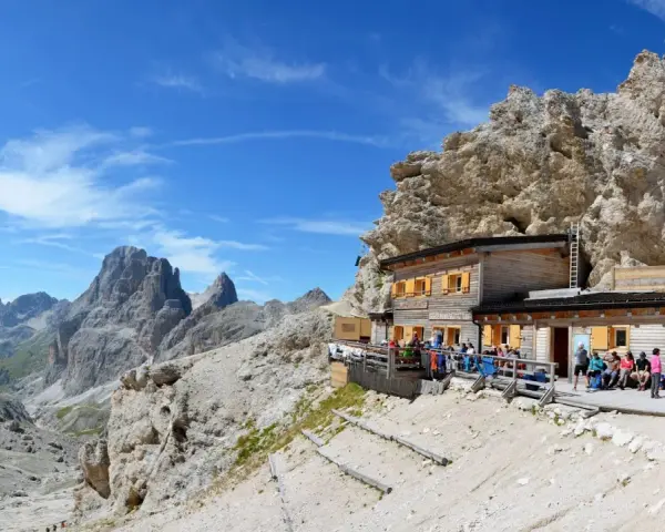 Rifugio alpino con gente seduta all'esterno, circondato da rocce imponenti e montagne. Un luogo ideale per le 12 escursioni da fare in Val di Fassa.