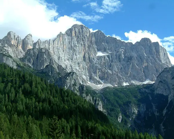 Imponente parete rocciosa del monte Civetta, con foreste di conifere ai suoi piedi e nuvole bianche nel cielo azzurro.