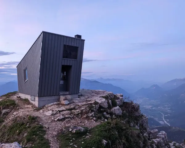 Il bivacco Giacomelli alla Madonnina, una struttura moderna su una cresta rocciosa, domina un paesaggio montano al tramonto.
