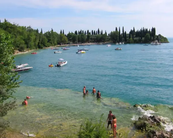 La baia delle Sirene a Garda, con gente che nuota e barche sull'acqua cristallina, sotto un cielo azzurro.