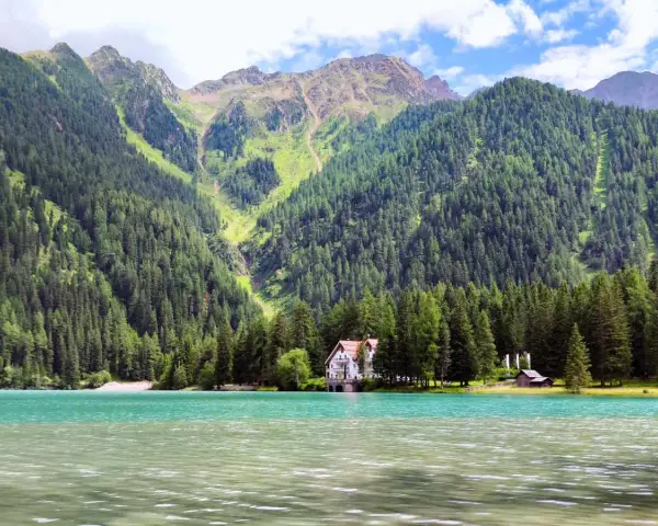 Un incantevole **Antholzer See** con acque turchesi, circondato da fitti boschi e imponenti montagne. Una casa bianca e un piccolo rifugio si affacciano sul lago.