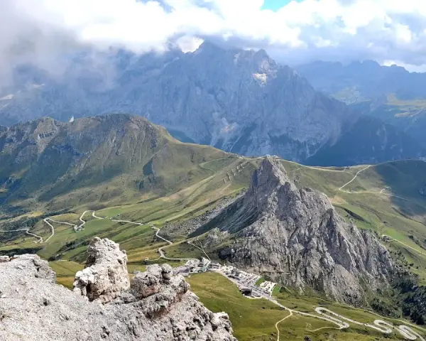 Panoramica mozzafiato del **Passo Pordoi** con strade tortuose che si snodano tra verdi pendii e imponenti vette rocciose, sotto un cielo nuvoloso.