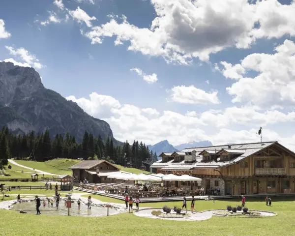 Un rifugio alpino con tavoli all'aperto e un laghetto dove giocano bambini, incorniciato da boschi e montagne imponenti sui **piani di Pezzè**.