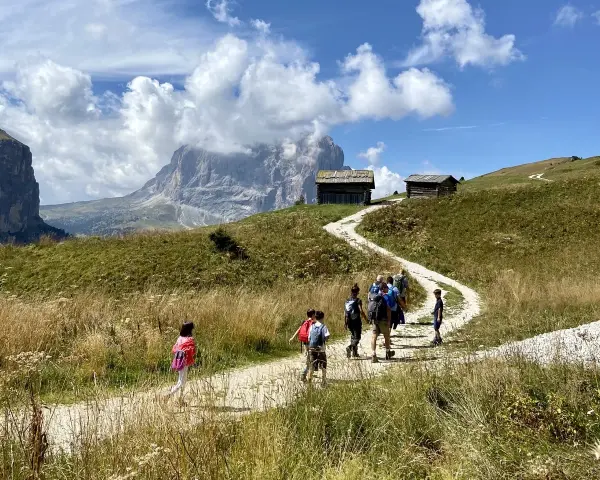 Famiglia in escursione su sentiero facile a Selva di Val Gardena, con baite e montagne sullo sfondo.