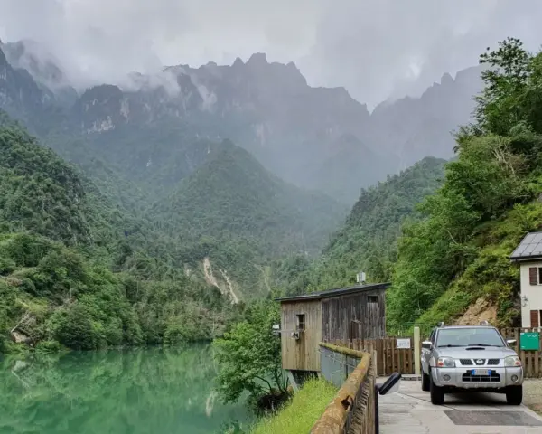 Il lago della Stua, incastonato tra montagne nebbiose e foreste lussureggianti, con una casa e un'auto parcheggiata vicino.