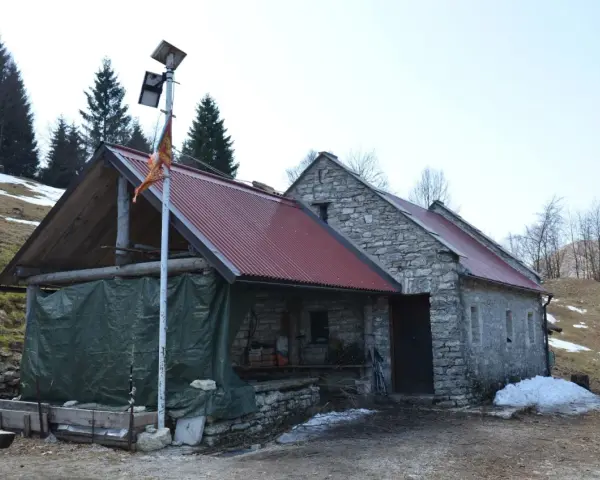 Bivacco Vallon Scuro: rifugio in pietra con tetto rosso, pannelli solari e telo verde. Un uomo si ripara dal freddo.