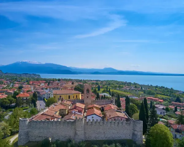 Vista aerea di Moniga del Garda, con il castello medievale in primo piano, il lago e le montagne sullo sfondo.