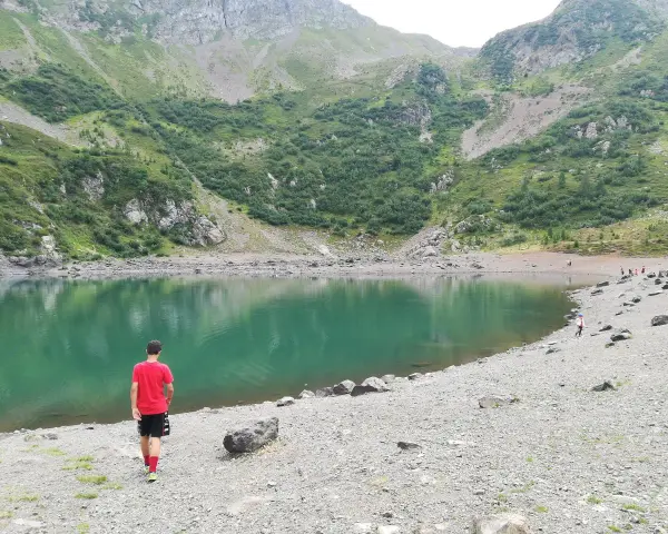 Ragazzo in maglietta rossa ammira il **lago di Erdemolo**, circondato da verdi pendii montuosi e rocce.