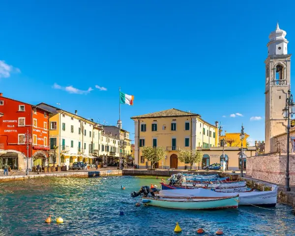 Un pittoresco porto dove si trova Lazise, con case colorate, barche e un campanile svettante sotto un cielo azzurro.