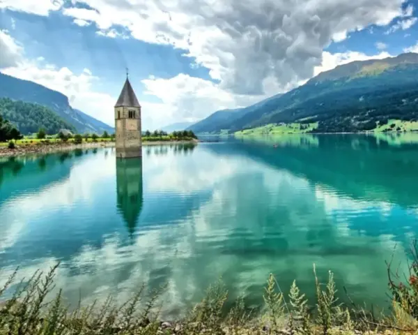 Torre sommersa nel lago di Resia, con montagne verdi e cielo nuvoloso.