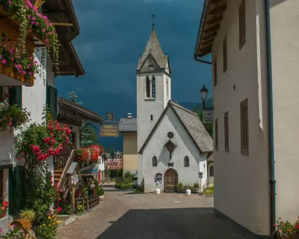 Un pittoresco vicolo a Rocca Pietore, con case fiorite e una chiesa dal cielo tempestoso.