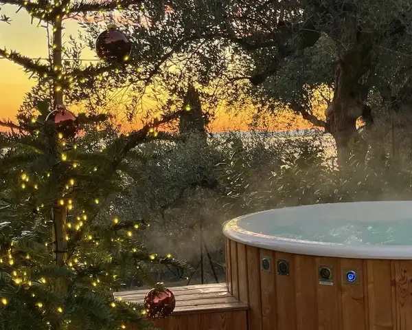 Un albero di Natale illuminato e una vasca idromassaggio fumante al tramonto sul Lago di Garda. Il camping è aperto tutto l'anno, un'oasi di relax invernale.