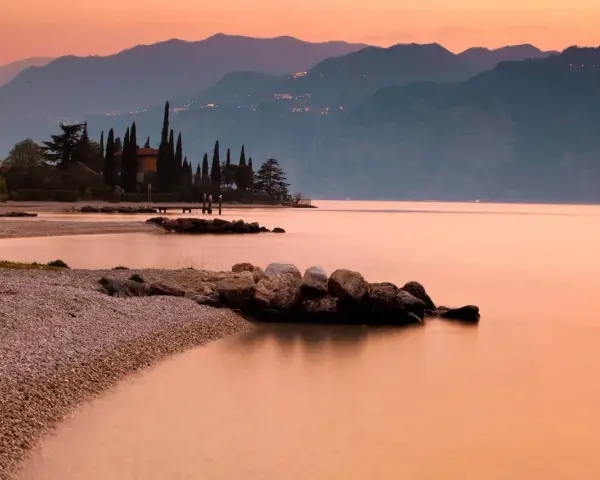 Brenzone sul Garda: cosa vedere? Spiaggia di ciottoli, cipressi e montagne al tramonto sul lago.