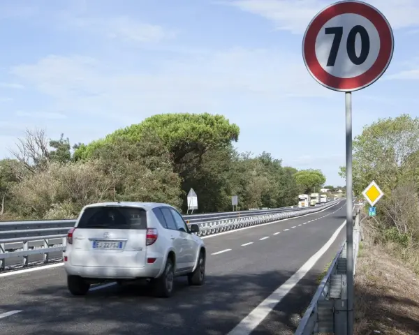 Un'auto bianca percorre una strada extraurbana con limite di 70 km/h. Alberi e cielo azzurro fanno da cornice.