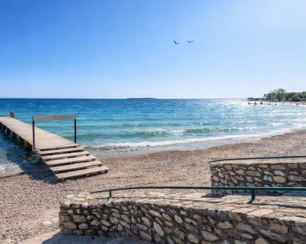Molo in legno sul **Manerba del Garda strand**, con acqua cristallina e cielo azzurro. Due gabbiani volano sopra il lago.