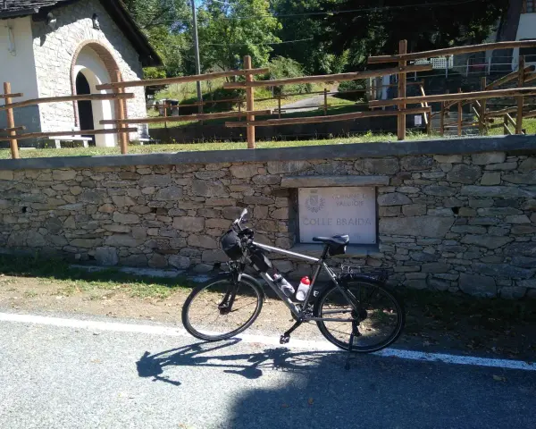 Bicicletta ferma sul ciglio della strada, con un muretto in pietra e un cartello che indica "Colle Braida".