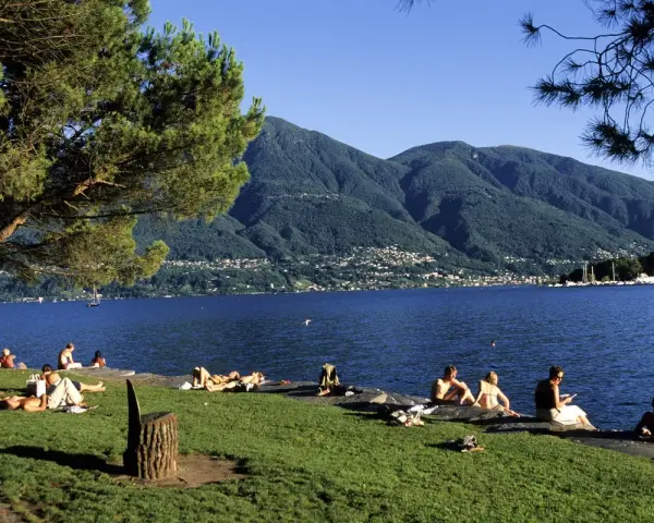 Persone rilassate sui prati e sulle rive di uno dei bellissimi laghi piemontesi, con montagne verdi sullo sfondo.