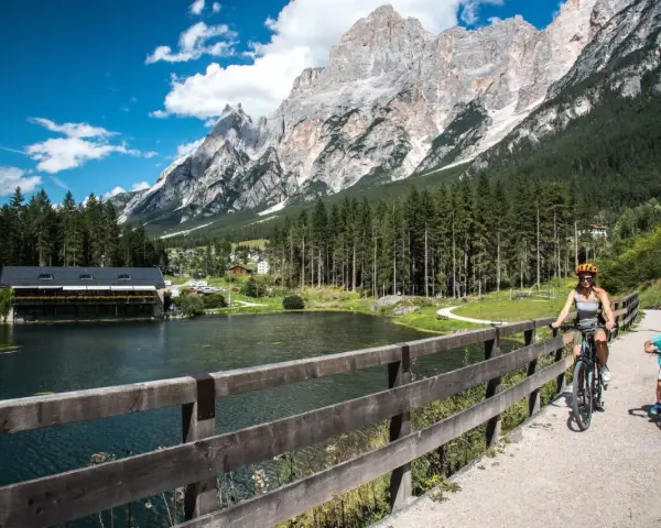 Famiglia in bicicletta sul ponte del **lago di San Vito**, con montagne imponenti e boschi sullo sfondo.