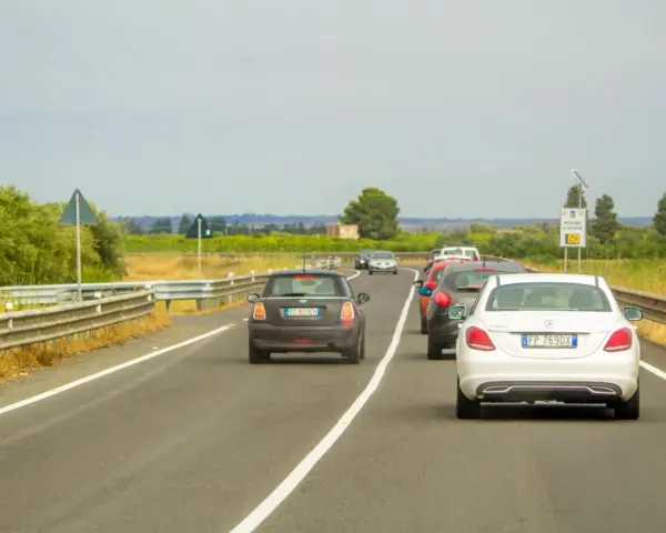 Traffico in colonna su strada con vegetazione ai lati. Un'auto tenta il sorpasso linea continua, mettendo a rischio la sicurezza.
