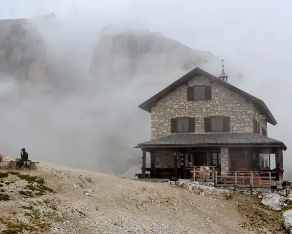 Rifugio Franz Kostner avvolto dalla nebbia, con un escursionista solitario su una panchina e le bandiere che sventolano.