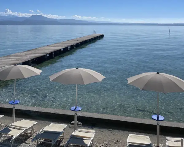 Ombrelloni e lettini al lido delle Bionde, con un pontile in legno che si estende sul lago limpido e montagne all'orizzonte.