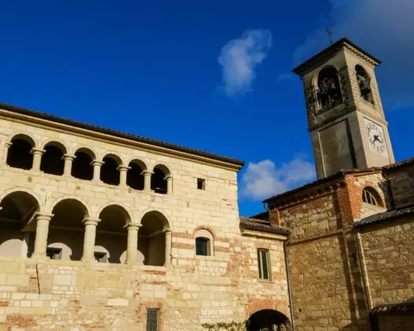 Antica **cella monte** con portici ad archi e torre campanaria sotto un cielo azzurro.