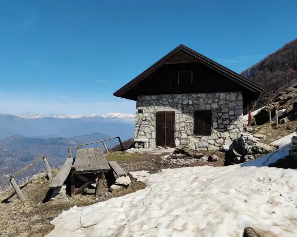 Bivacco Monte Grappa, rifugio in pietra con tavolo da picnic e vista sulle montagne innevate.
