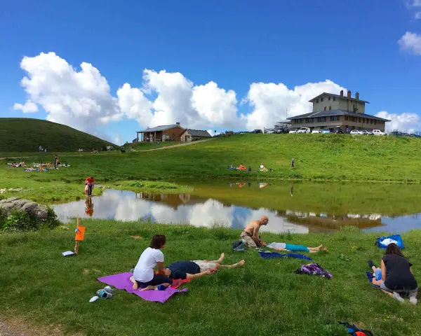 Persone rilassate sui prati verdi, vicino a un lago, sotto un cielo azzurro con nuvole bianche. Sembra un luogo ideale per i piani dell'avaro, godendosi la natura.