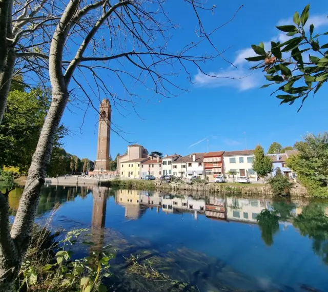 Un tranquillo fiume riflette le case colorate e l'alto campanile di Casale sul Sile, con alberi che incorniciano la scena.