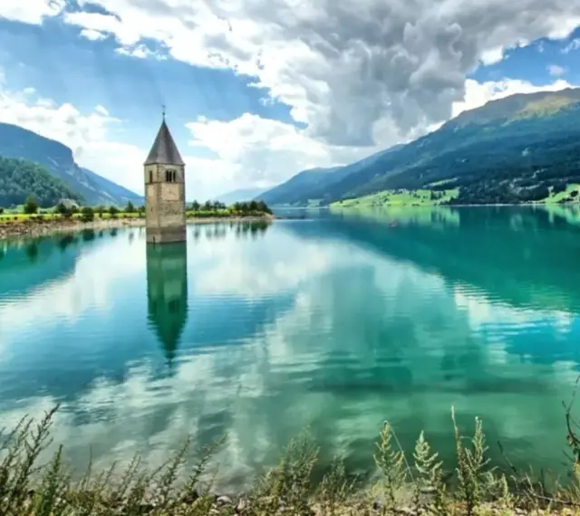 Torre sommersa nel lago di Resia, con montagne verdi e cielo nuvoloso.