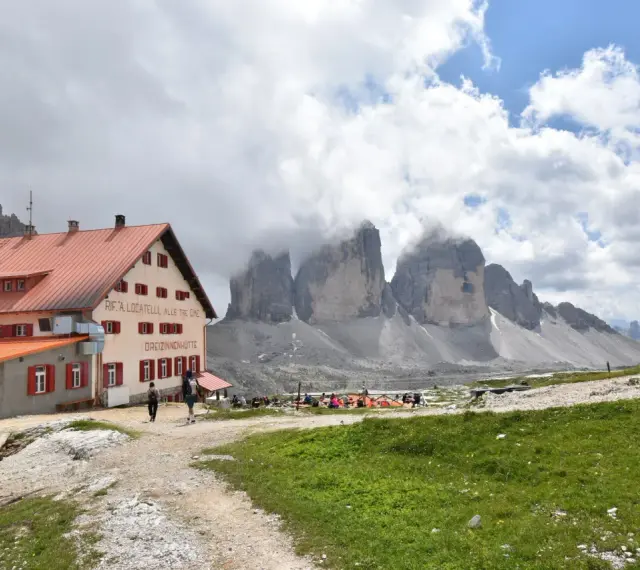 Il rifugio Locatelli, con le Tre Cime di Lavaredo avvolte dalle nuvole, accoglie escursionisti su un prato verde.