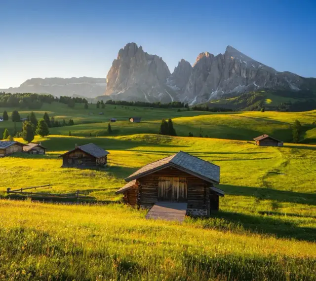 Alpe di Siusi al tramonto, con baite in legno su prati verdi e montagne imponenti sullo sfondo.