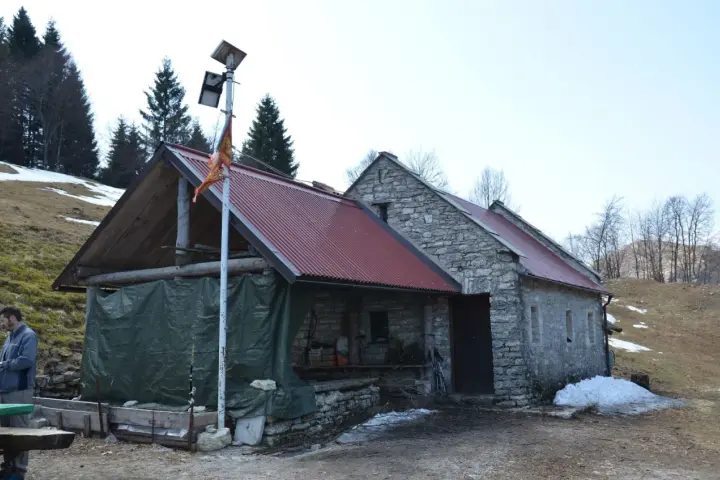 Bivacco Vallon Scuro: rifugio in pietra con tetto rosso, pannelli solari e telo verde. Un uomo si ripara dal freddo.