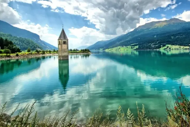 Torre sommersa nel lago di Resia, con montagne verdi e cielo nuvoloso.