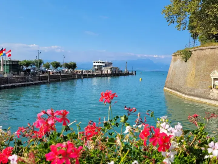 Fiori rossi e bianchi in primo piano, con un molo, un traghetto e montagne sullo sfondo. Un'immagine che evoca i paesi vicino Peschiera del Garda.