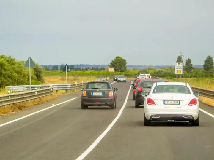Traffico in colonna su strada con vegetazione ai lati. Un'auto tenta il sorpasso linea continua, mettendo a rischio la sicurezza.