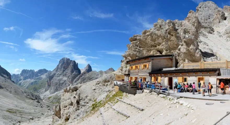 Rifugio alpino con gente seduta all'esterno, circondato da rocce imponenti e montagne. Un luogo ideale per le 12 escursioni da fare in Val di Fassa.