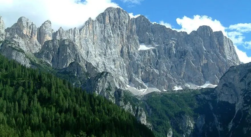 Imponente parete rocciosa del monte Civetta, con foreste di conifere ai suoi piedi e nuvole bianche nel cielo azzurro.