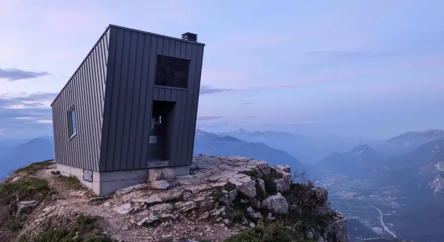 Il bivacco Giacomelli alla Madonnina, una struttura moderna su una cresta rocciosa, domina un paesaggio montano al tramonto.