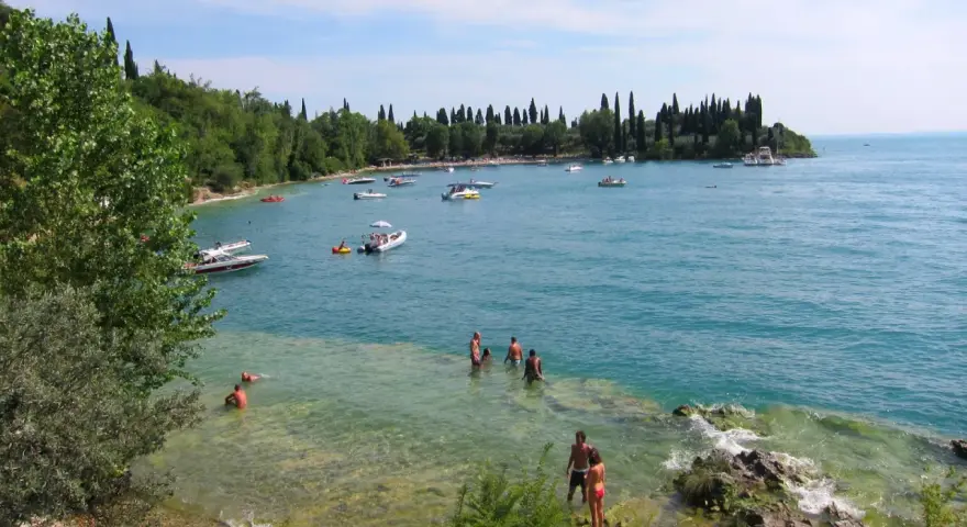 La baia delle Sirene a Garda, con gente che nuota e barche sull'acqua cristallina, sotto un cielo azzurro.