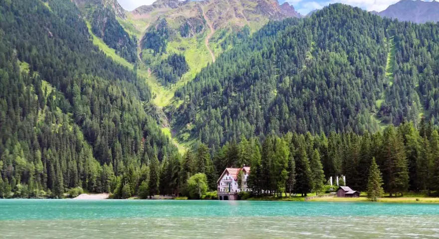 Un incantevole **Antholzer See** con acque turchesi, circondato da fitti boschi e imponenti montagne. Una casa bianca e un piccolo rifugio si affacciano sul lago.