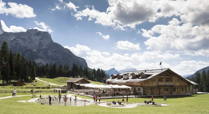 Un rifugio alpino con tavoli all'aperto e un laghetto dove giocano bambini, incorniciato da boschi e montagne imponenti sui **piani di Pezzè**.