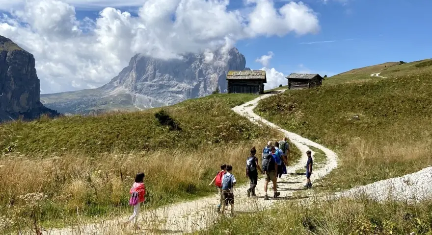 Famiglia in escursione su sentiero facile a Selva di Val Gardena, con baite e montagne sullo sfondo.