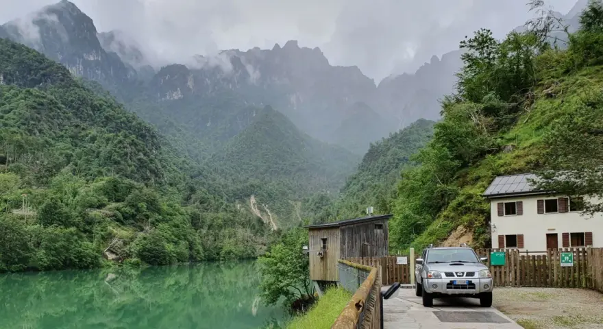 Il lago della Stua, incastonato tra montagne nebbiose e foreste lussureggianti, con una casa e un'auto parcheggiata vicino.