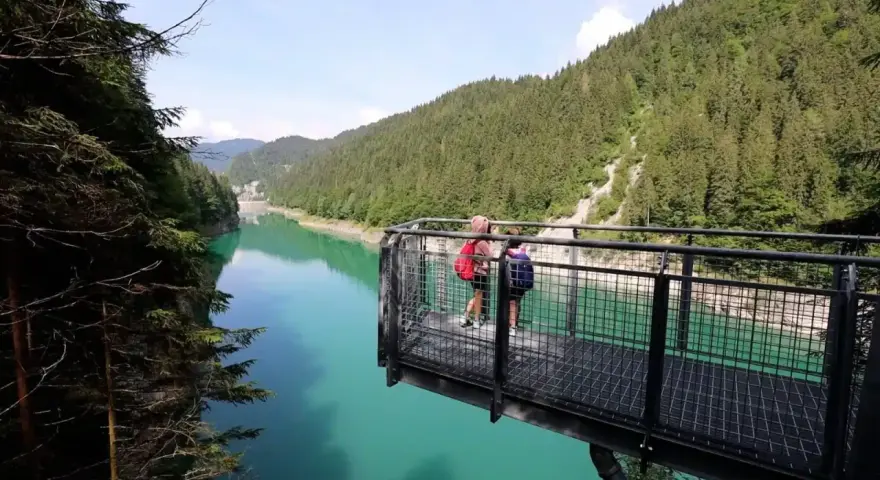 Due persone su una piattaforma panoramica ammirano il lago di Val Noana. Ecco come arrivare a questo splendido lago alpino.