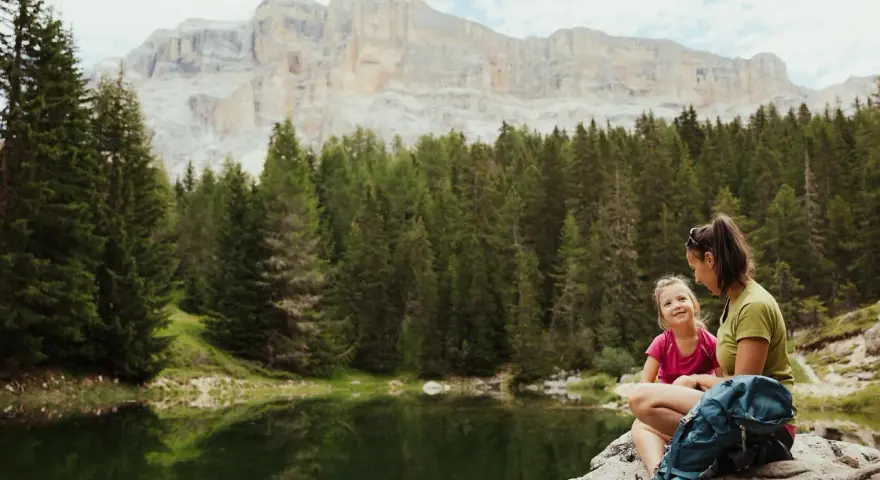 Mamma e figlia sedute su una roccia vicino a un lago alpino, con uno zaino blu. Un momento di relax per il loro lech dla lunch, con montagne imponenti sullo sfondo.