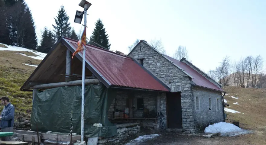 Bivacco Vallon Scuro: rifugio in pietra con tetto rosso, pannelli solari e telo verde. Un uomo si ripara dal freddo.