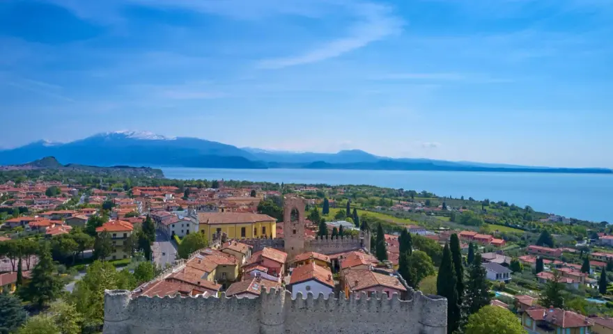 Vista aerea di Moniga del Garda, con il castello medievale in primo piano, il lago e le montagne sullo sfondo.