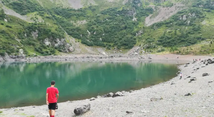 Ragazzo in maglietta rossa ammira il **lago di Erdemolo**, circondato da verdi pendii montuosi e rocce.