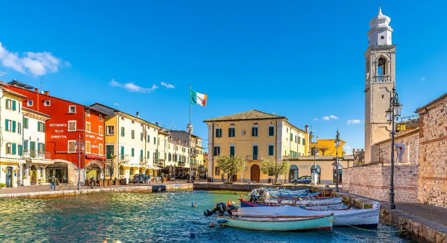 Un pittoresco porto dove si trova Lazise, con case colorate, barche e un campanile svettante sotto un cielo azzurro.